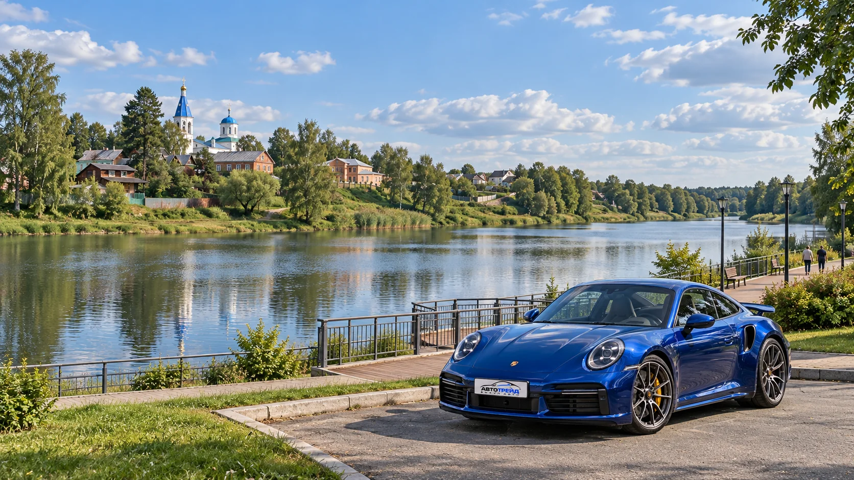 Синий Porsche 911 Turbo S с логотипом Автотрейд у водоёма Городня в Волоколамске, на фоне зелёного берега и храма